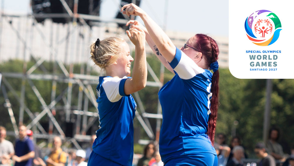 Kaksi iloista SO-tyttö-jalkapalloilijaa tuulettamassa kentällä. Kuvan oikeassa yläkulmassa valkoisella taustalla Santiago 2027 kisalogo.